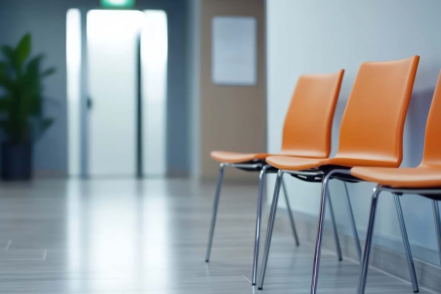 A row of empty chairs stands in an office waiting area. The chairs are a bright orange color and have sleek, modern lines with silver metal legs. The room has a bright, neutral color palette with a light wood floor. A plant can be seen in the background, adding a touch of nature to the space. The scene evokes a sense of calm and order. This image would work well for concepts related to business, healthcare, waiting, or interior design. The image also has copy space. Credit: Vecteezy